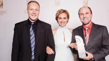 Jens Baumgart und Patrik Buchmüller (RPR1), Preisträger in der Kategorie "Bestes Nachrichtenformat", mit Laudatorin Caren Miosga vor einer Fotowand im Schuppen 52 in Hamburg. © NDR 