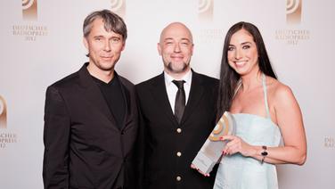 Bettina Zacher und Stephan Heller (Klassik Radio), Preisträger in der Kategorie "Beste Programmaktion", posiert mit Laudator Der Graf von Unheilig vor einer Fotowand im Schuppen 52 in Hamburg. © NDR 