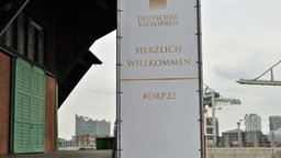 Neben dem Schuppen 52 im Hamburger Hafen steht ein Banner mit der Aufschrift "Deutscher Radiopreis 2022", im Hintergrund die Elbphilharmonie und Kräne © Deutscher Radiopreis / Janine Kühl Foto: Janine Kühl